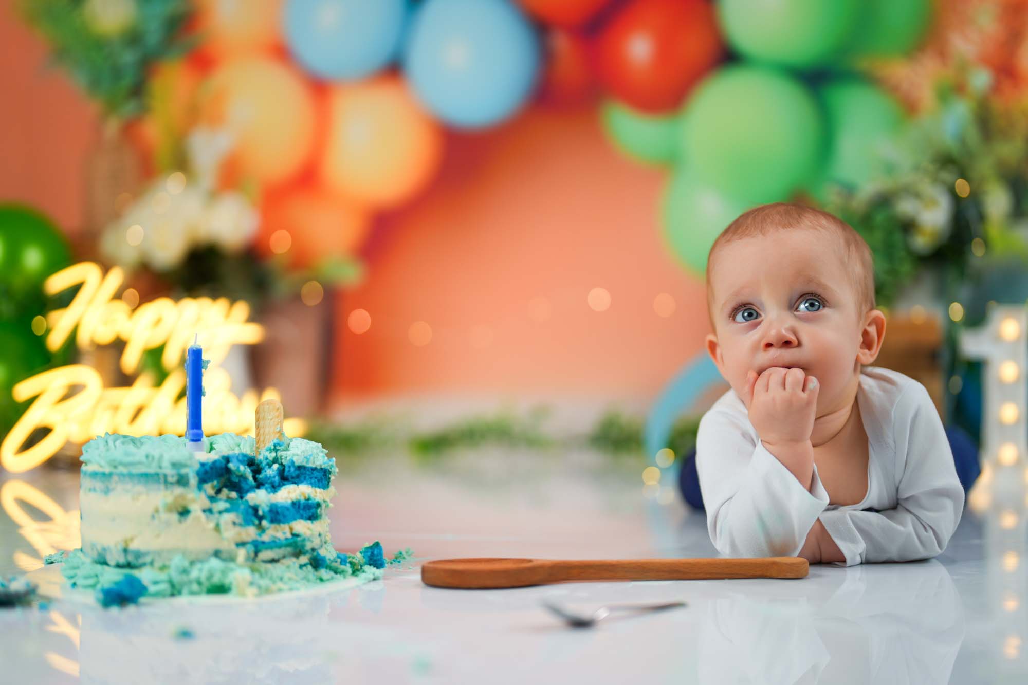 Ein Baby-Junge mit neugierigem Blick sitzt vor seiner blauen Geburtstagstorte während eines Cake-Smash-Shootings.
