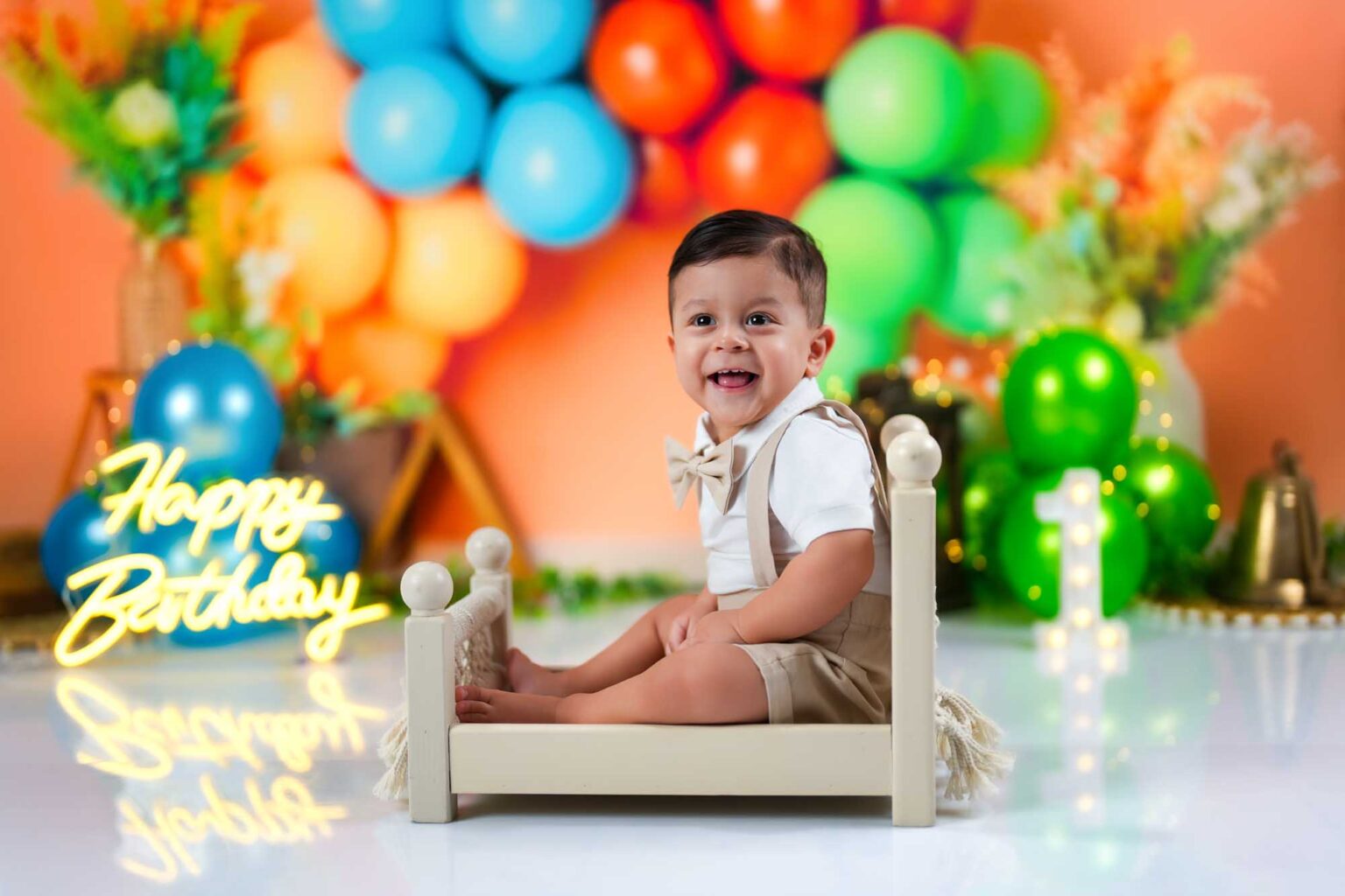Lächelnder Baby-Junge in festlicher Kleidung sitzt auf einem kleinen Bett vor bunten Ballons und einem Happy-Birthday-Neonlicht.