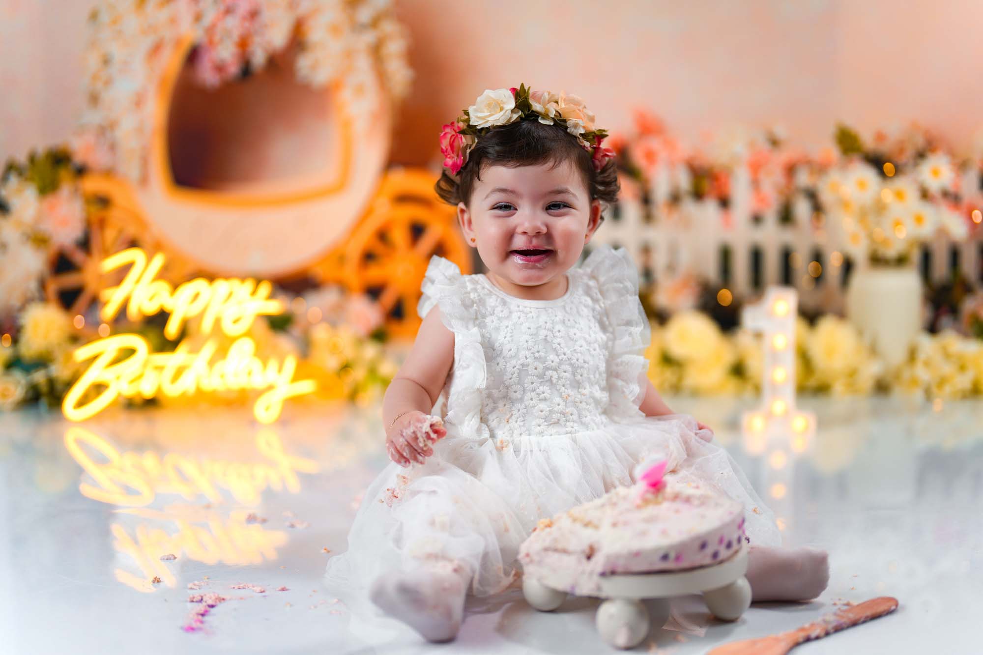 Lächelndes Baby-Mädchen in einem weißen Kleid mit Blumenkranz vor einem Happy-Birthday-Neonlicht.