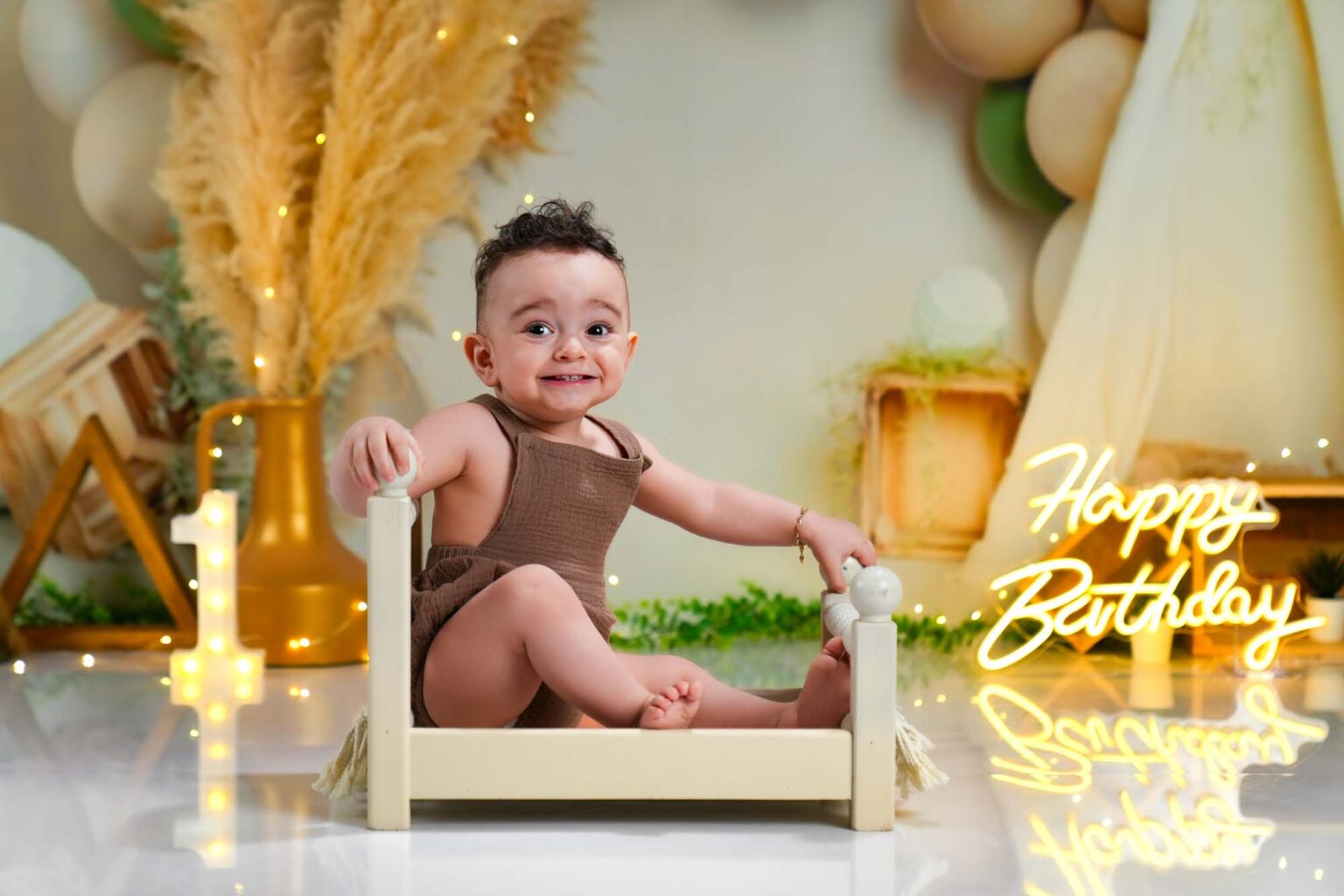 Baby im braunen Outfit sitzt lachend auf einem kleinen Holzbett.