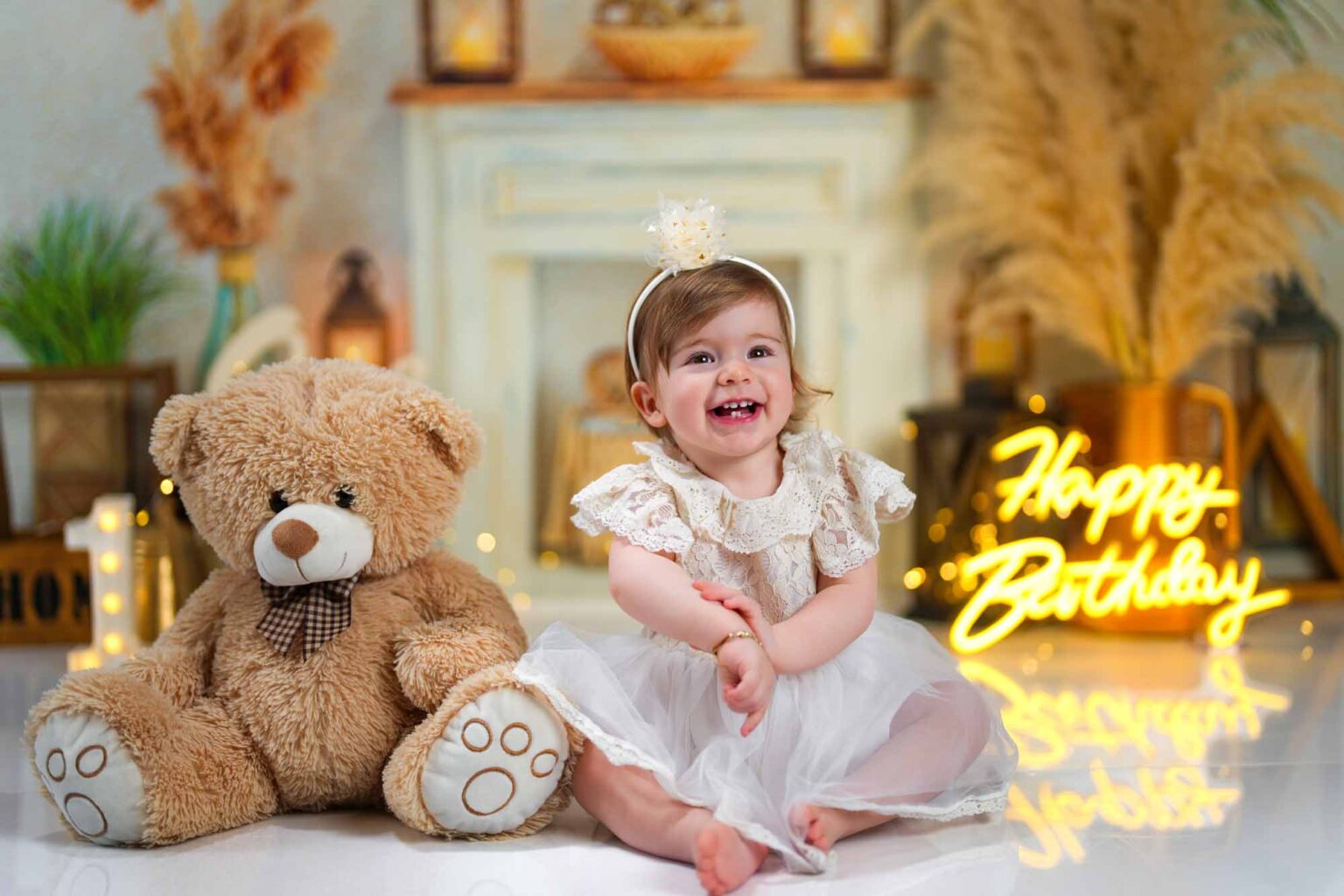 Ein lachendes Baby im weißen Spitzenkleid sitzt neben einem großen Teddybären vor einer gemütlichen Geburtstagskulisse.