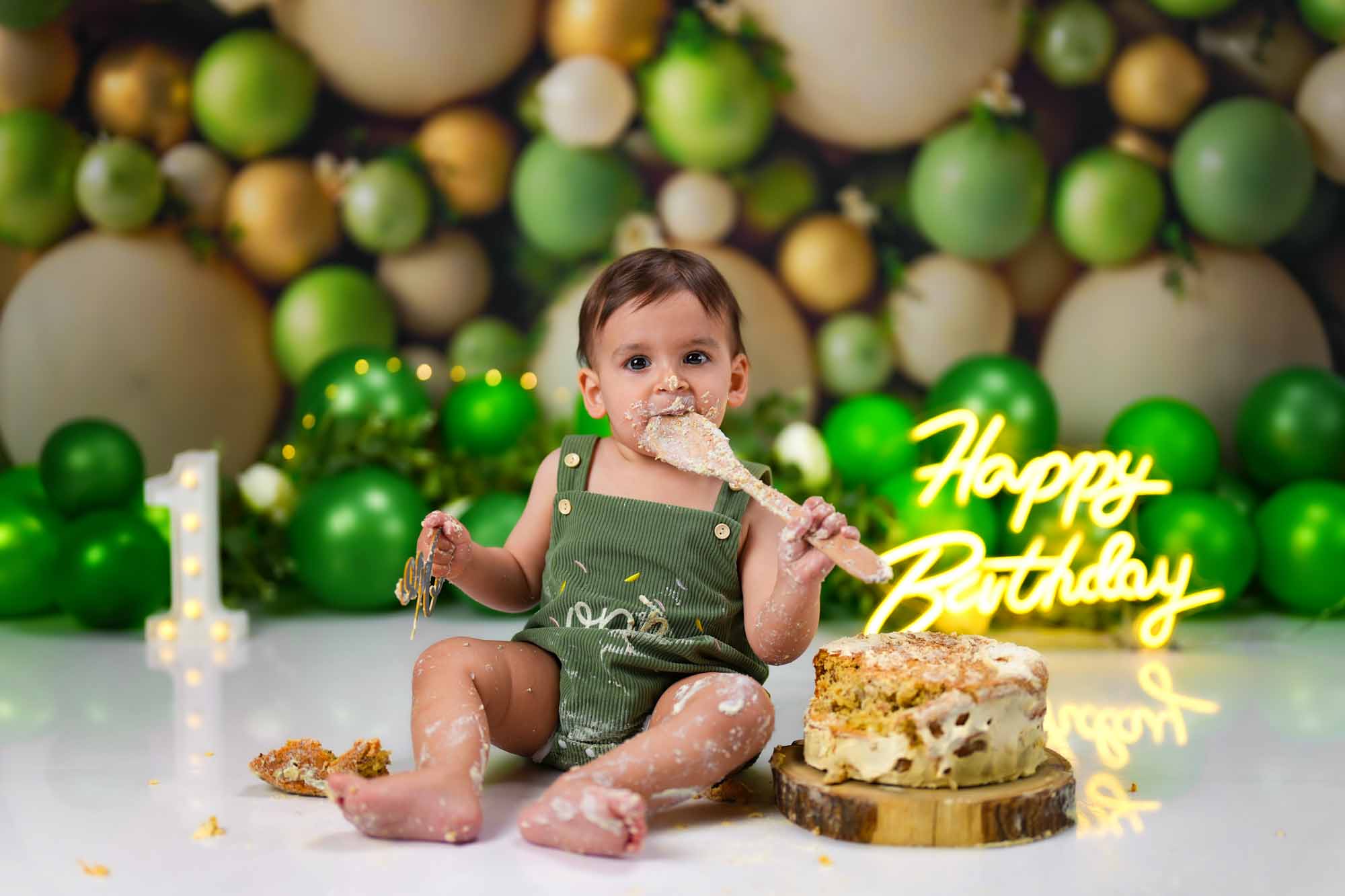Baby im grünen Outfit beim Cake Smash Shooting mit grün-goldener Ballondekoration und „Happy Birthday“-Leuchtschrift.