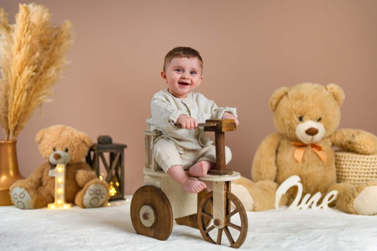 Baby in cremefarbenem Outfit auf einem Vintage-Holzdreirad mit Teddybären und Geburtstagsdeko.