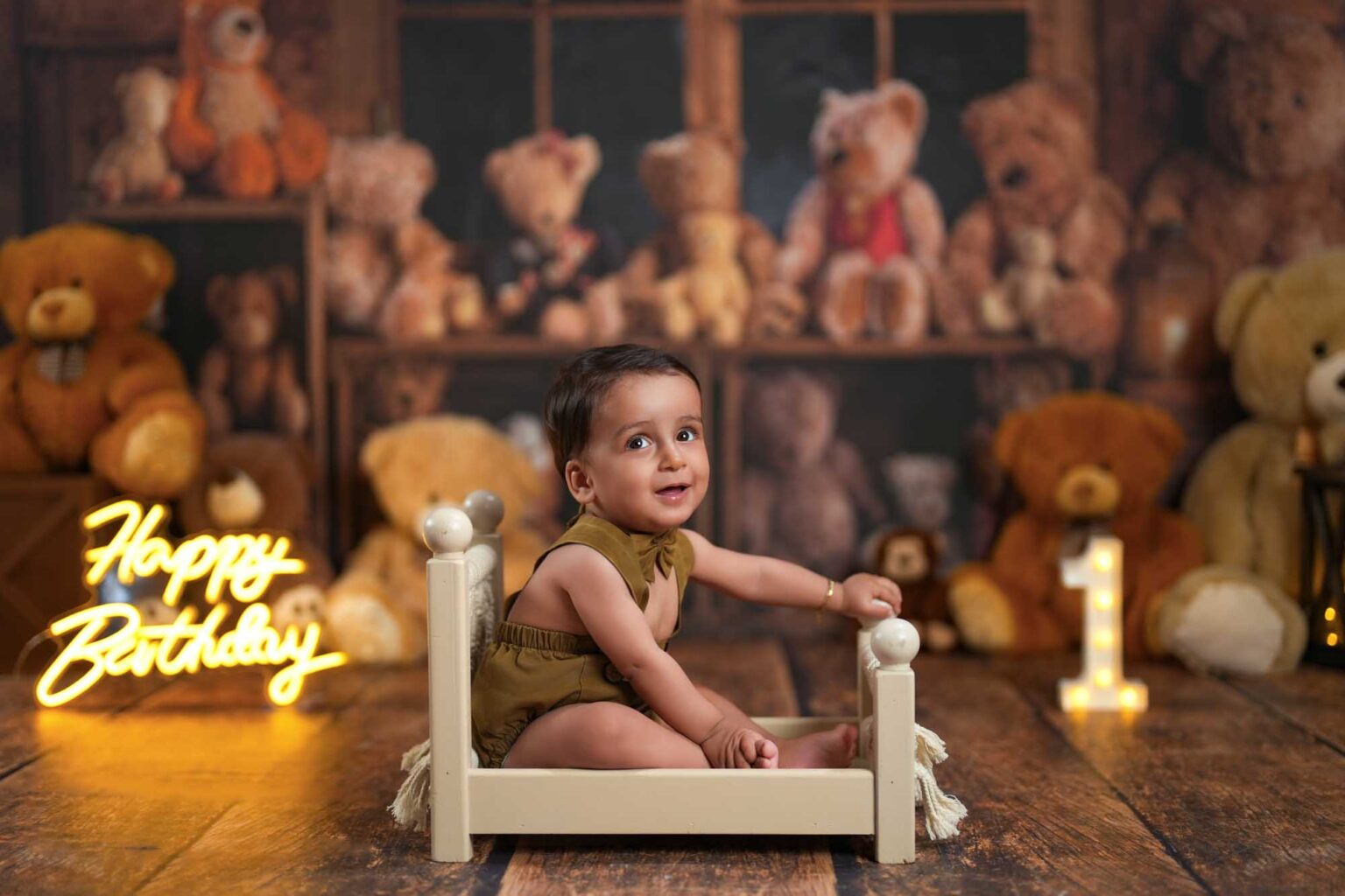Baby sitzt lachend in einem kleinen cremefarbenen Holzbett vor einer Kulisse voller Plüsch-Teddybären.
