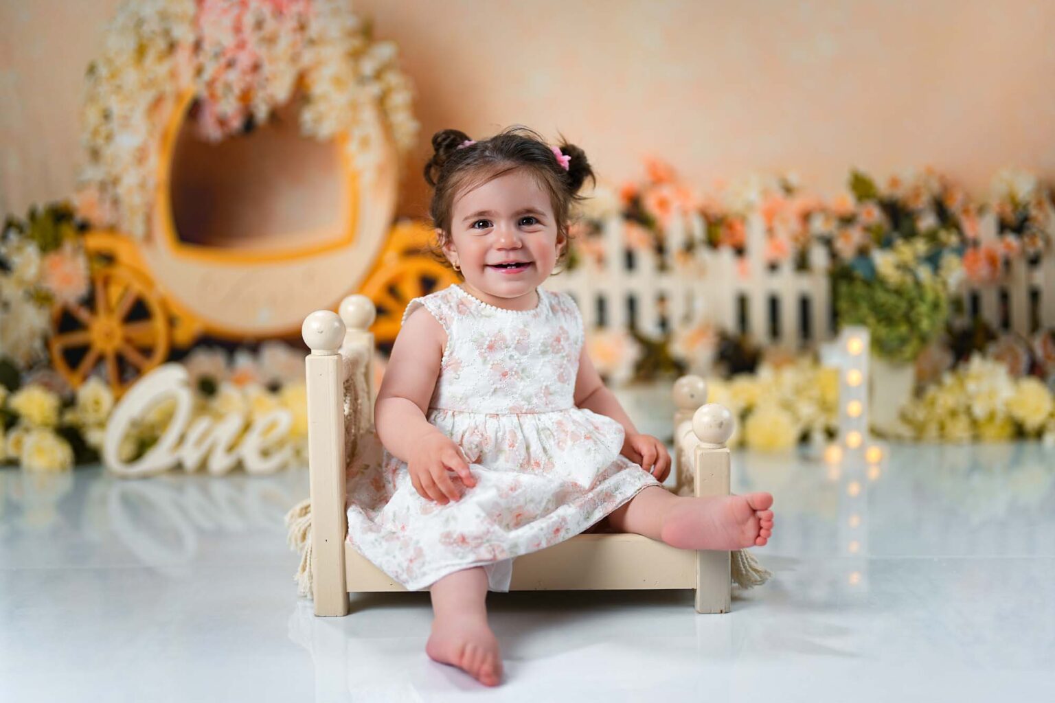 Baby im weißen Blumenkleid sitzt lächelnd im kleinen Bett mit romantischer Blumenkulisse.