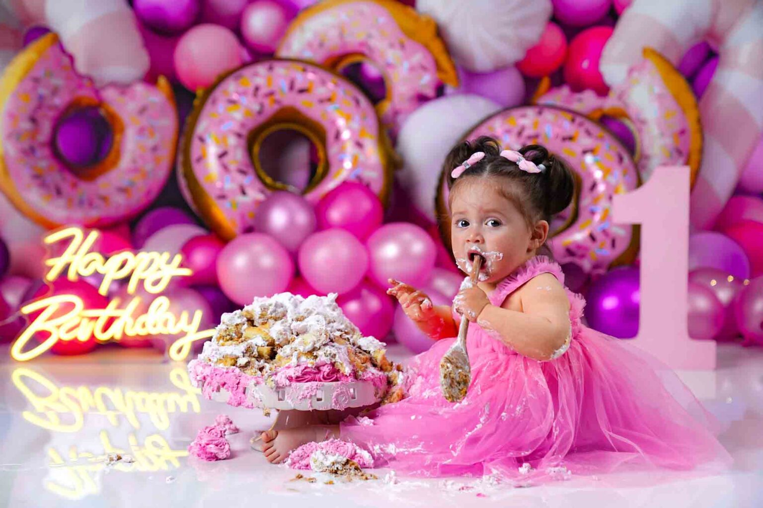 Mädchen im pinken Tüllkleid macht ein Cake Smash Shooting vor einer bunten Donut- und Ballonwand