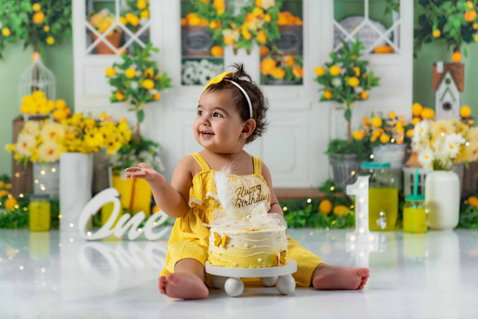 Ein lächelndes Baby-Mädchen in einem gelben Outfit feiert ihren ersten Geburtstag mit einer gelben Torte und 'One'-Schild.