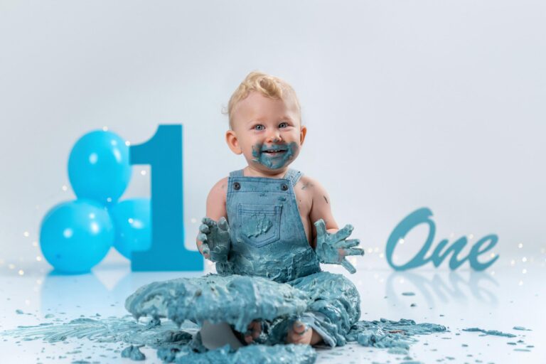 Baby in blauer Latzhose sitzt vor einer blauen Geburtstagstorte und genießt den Cake Smash.
