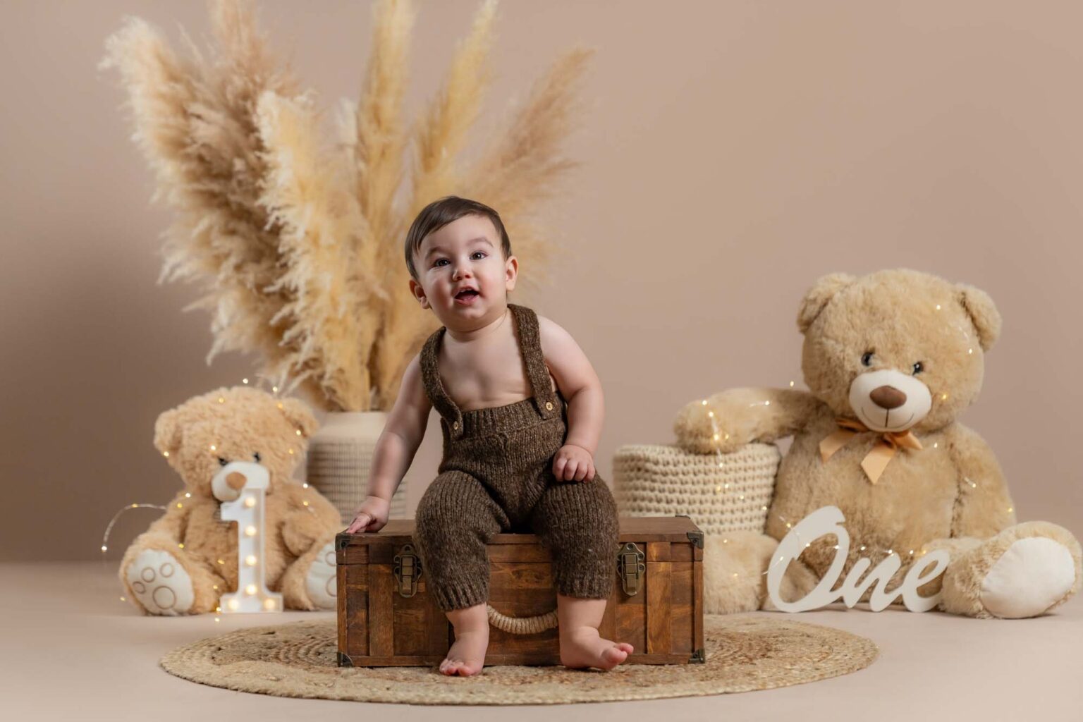 Baby im braunen Strickoverall sitzt auf einer Holztruhe vor Teddy-Dekoration zum 1. Geburtstag.