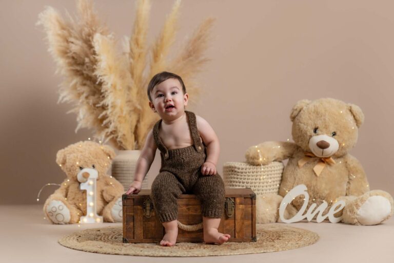 Baby im braunen Strickoverall sitzt auf einer Holztruhe vor Teddy-Dekoration zum 1. Geburtstag.