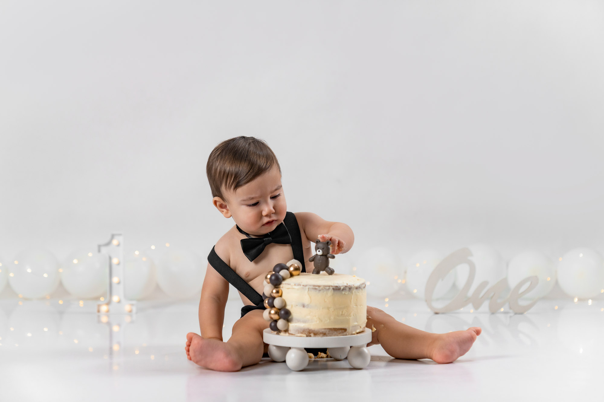 Baby im schwarzen Hosenträger-Set spielt mit zerdrückter Geburtstagstorte auf weißem Boden.