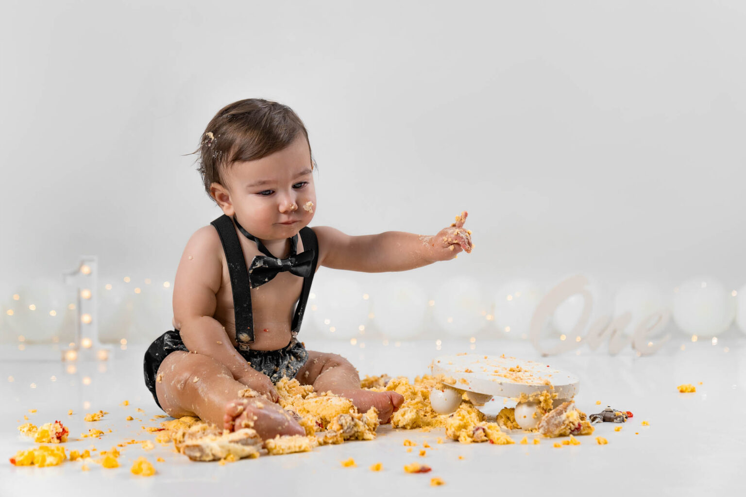 Baby im schwarzen Hosenträger-Set mit Fliege sitzt vor einer Torte, minimalistisch dekoriert mit weißen Luftballons im Hintergrund.