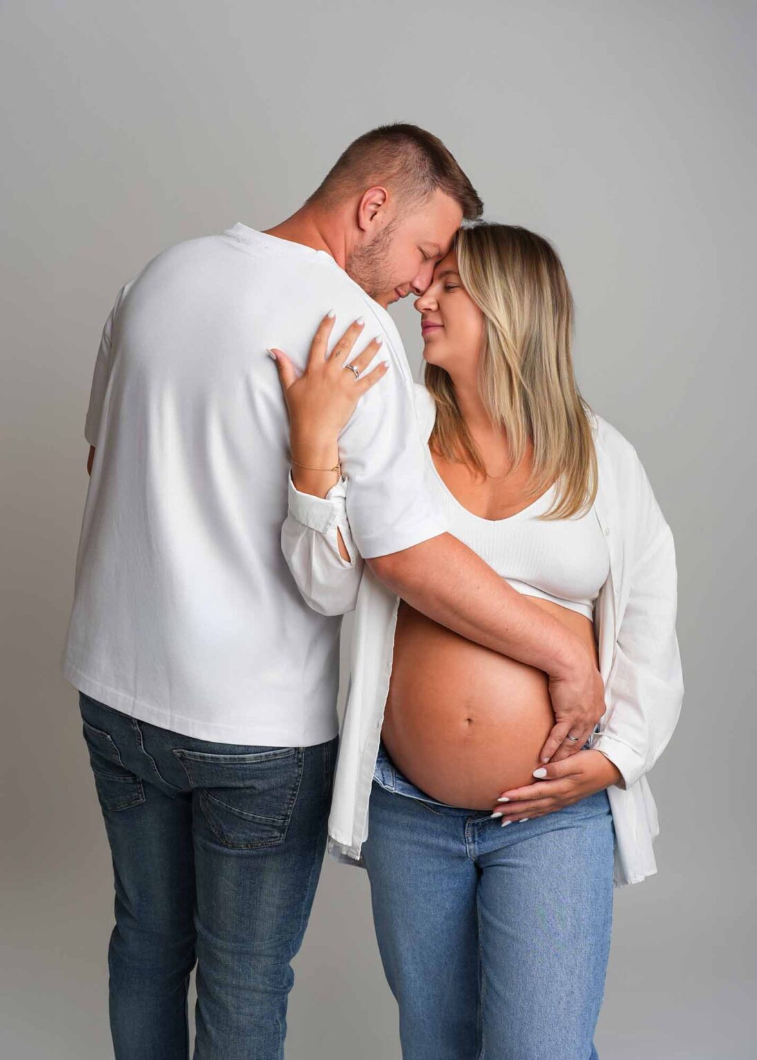 Ein werdendes Paar in weißen Outfits steht in einem Studio und umarmt zärtlich den Babybauch der Frau.
