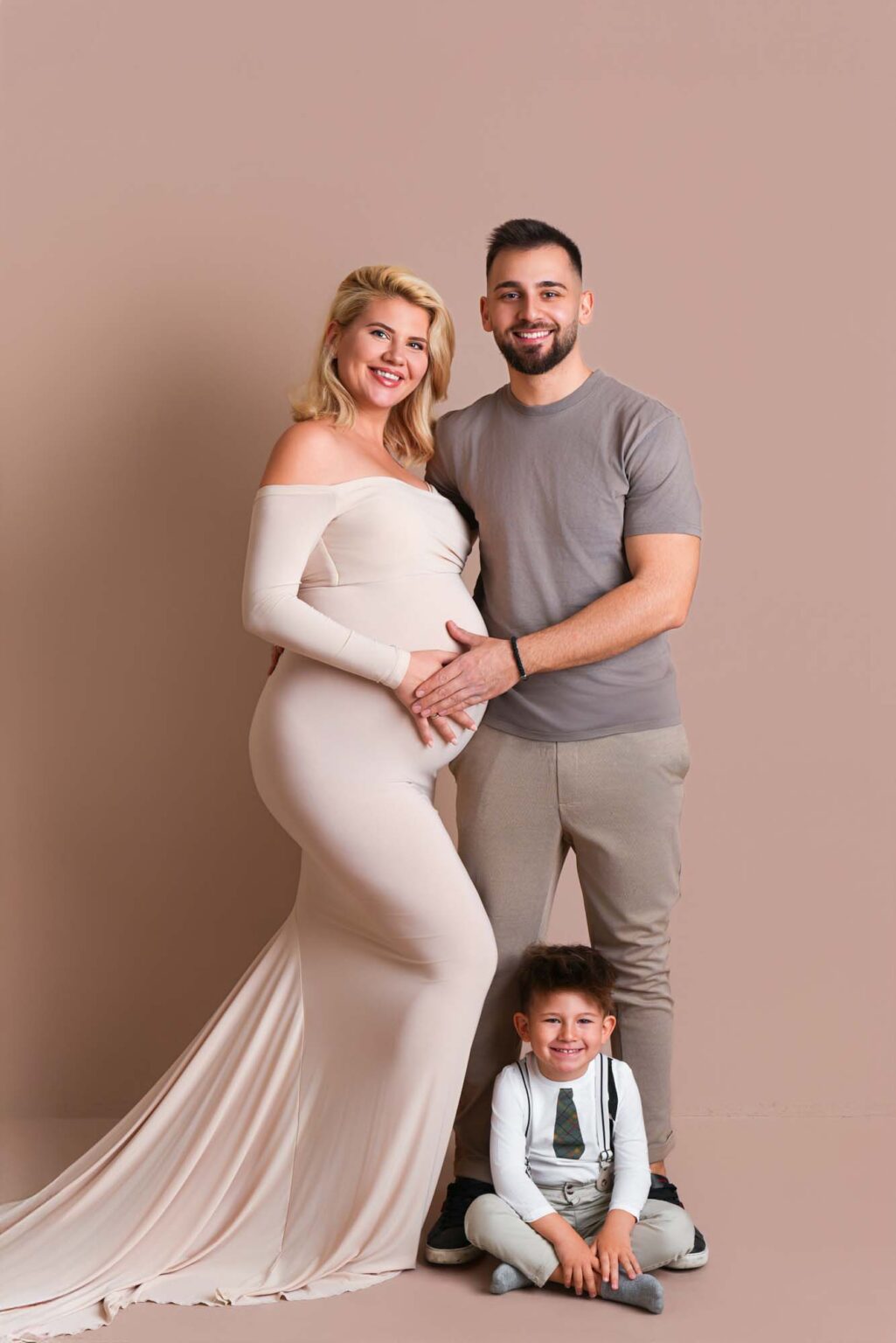 Eine lächelnde werdende Familie in einem Studio-Porträt, mit der werdenden Mutter in einem eleganten weißen Kleid und ihrem Kind davo.